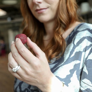 May include: A woman's hand holding a small, round, red candy. The candy is smooth and has a slight sheen. The woman is wearing a ring on her finger.