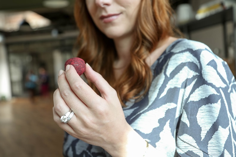 May include: A woman's hand holding a small, round, red candy. The candy is smooth and has a slight sheen. The woman is wearing a ring on her finger.