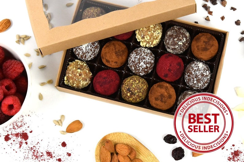 May include: A box of assorted vegan truffles in a variety of flavors and colors. The box is open and the truffles are arranged in a grid pattern. The box is labeled "Delicious Indecisions" and "Best Seller".