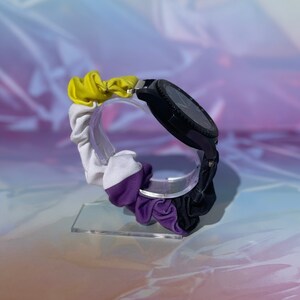 May include: A black smartwatch with a yellow, white, and purple fabric band. The band is a scrunchie style and is on a clear display stand.