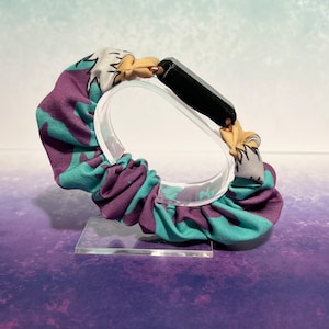 May include: A purple and teal fabric scrunchie with a white and yellow design. The scrunchie is on a clear display stand.