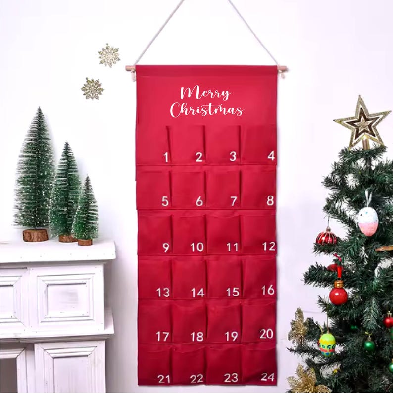 May include: Red fabric Christmas advent calendar with 24 numbered pockets.  The calendar features the text 'Merry Christmas' in white script lettering at the top.  It hangs from a simple wooden dowel and string. The pockets are numbered 1-24.  A festive Christmas tree and miniature pine trees are visible in the background.