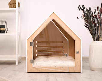 May include: A white and wood dog house with a clear acrylic door and a white cushion inside. The house is on a light wood floor.
