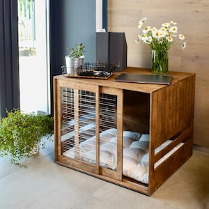 May include: A wooden dog crate with a sliding door and a cushioned bed inside. The crate is brown with a natural wood finish and has a metal grid on the front. The crate is sitting on a wooden floor.