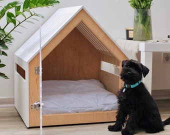 May include: A white wooden dog house with a gray cushion inside. A black dog with a turquoise collar sits in front of the house.