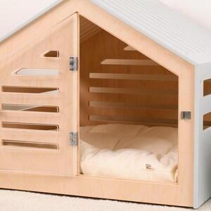 May include: A pet house featuring a light wood frame and a light gray roof and side panels. The front door is made of light wood with horizontal slats and a metal latch. Inside, a soft, cream-colored cushion covers the floor of the structure.