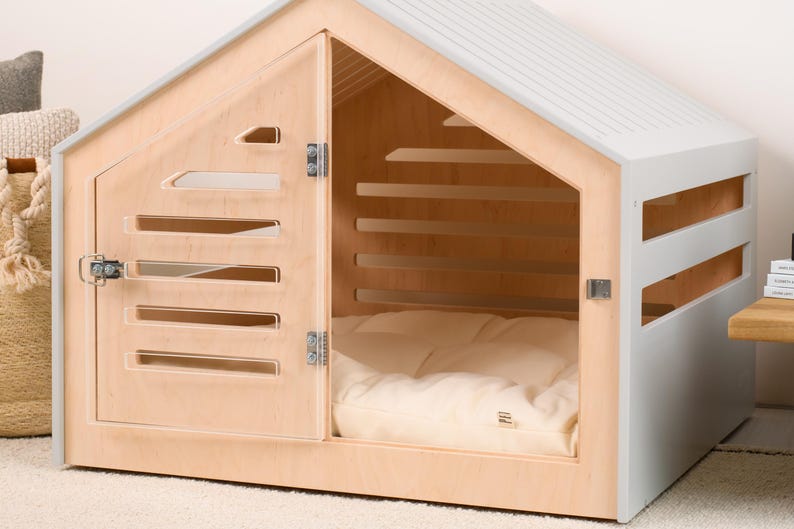 May include: A pet house featuring a light wood frame and a light gray roof and side panels. The front door is made of light wood with horizontal slats and a metal latch. Inside, a soft, cream-colored cushion covers the floor of the structure.