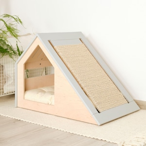 May include: A wooden cat house with a gray roof and a sisal scratching ramp. The house has a light-colored wood frame and a soft, cream-colored cushion inside. The ramp is angled and provides a surface for cats to scratch.