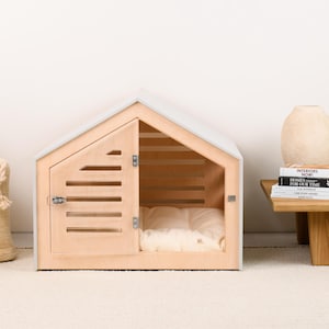 May include: A light-colored wooden pet house with a gabled roof, featuring a hinged door with horizontal slats and a metal latch. Inside the structure, a soft, cream-colored cushioned bed is visible, providing a comfortable space for a small animal.