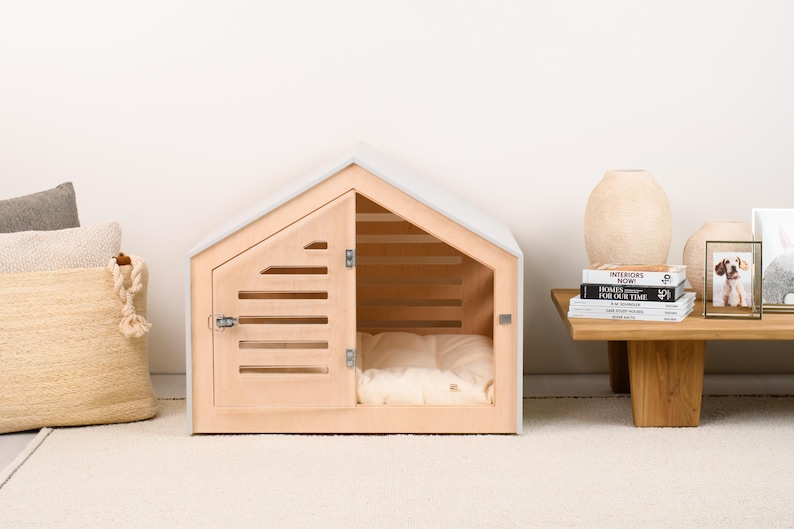 May include: A light-colored wooden pet house with a gabled roof, featuring a hinged door with horizontal slats and a metal latch. Inside the structure, a soft, cream-colored cushioned bed is visible, providing a comfortable space for a small animal.