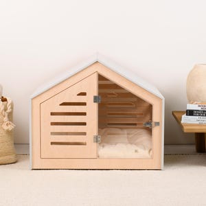 May include: A light-colored wooden pet house with a gabled roof, featuring a hinged door with horizontal slats and a clear acrylic panel. Inside, a soft, cream-colored cushion is visible. The structure has a light gray frame and metal hinges.
