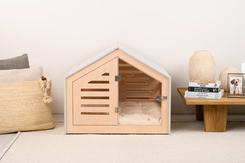 May include: A light-colored wooden pet house with a gabled roof, featuring a hinged door with horizontal slats and a clear acrylic panel. Inside, a soft, cream-colored cushion is visible. The structure has a light gray frame and metal hinges.