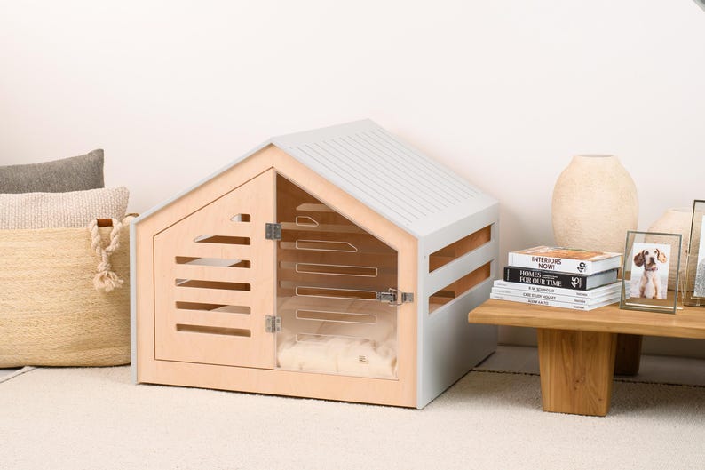 May include: A light gray and natural wood pet house with a gabled roof, featuring a front door with horizontal slats and a clear acrylic panel. The structure includes side ventilation slats and a metal latch on the door, with a soft white cushion inside.