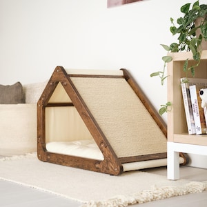 May include: A wooden cat house with a ramp and a bed. The frame is dark brown, with a woven sisal ramp and a cream-colored bed and interior. The cat house is placed on a cream-colored rug.