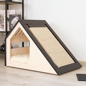 May include: A modern cat house with a light wood frame and a dark brown roof and scratching ramp. The house has a cozy interior with a cream-colored cushion. The ramp is covered in a woven sisal material.