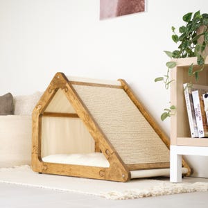 May include: A wooden cat house with a ramp and bed. The frame is made of light brown wood, with a woven sisal ramp and a cream-colored bed inside. The house is on a white rug.
