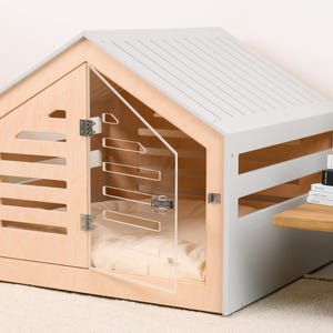 May include: A pet crate with a light wood frame and a light gray roof. The structure features horizontal slats on the sides and a clear acrylic door with metal hinges and a latch. Inside, a soft, light-colored cushion is visible for comfort.