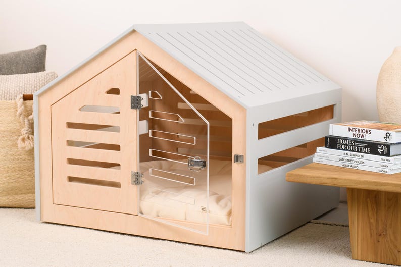 May include: A pet crate with a light wood frame and a light gray roof. The structure features horizontal slats on the sides and a clear acrylic door with metal hinges and a latch. Inside, a soft, light-colored cushion is visible for comfort.
