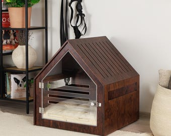 May include: A wooden dog house with a house-shaped design. The dark brown structure features a clear acrylic front door and a slatted roof. A white dog bed is visible inside. The dog house sits on a cream-colored rug.