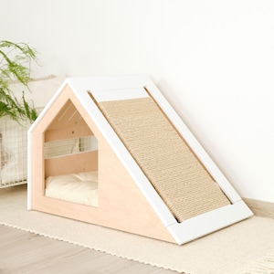 May include: A modern cat house with a light wood frame and white trim. It features a built-in ramp with a woven sisal scratching surface and a cozy interior with a cream-colored cushion. The house is designed in a minimalist style.