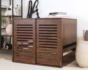 May include: A wooden dog crate with a dark brown finish. The crate features sliding doors with horizontal slats for ventilation. The design includes a pull-out tray and is placed on a white rug. A black harness hangs above the crate.