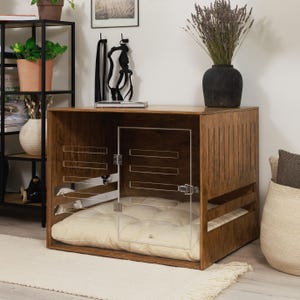 May include: A wooden dog crate with a clear acrylic door and a cream-colored cushion inside. The crate has a rectangular shape with horizontal slats on the sides and a solid top. The wood has a warm, brown finish. The crate is designed for indoor use.