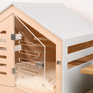 May include: A pet house featuring a light wood frame and a light gray gabled roof. The structure includes horizontal side slats and a clear acrylic door with a metal latch. Inside, a soft cream-colored cushion is visible through the transparent door.