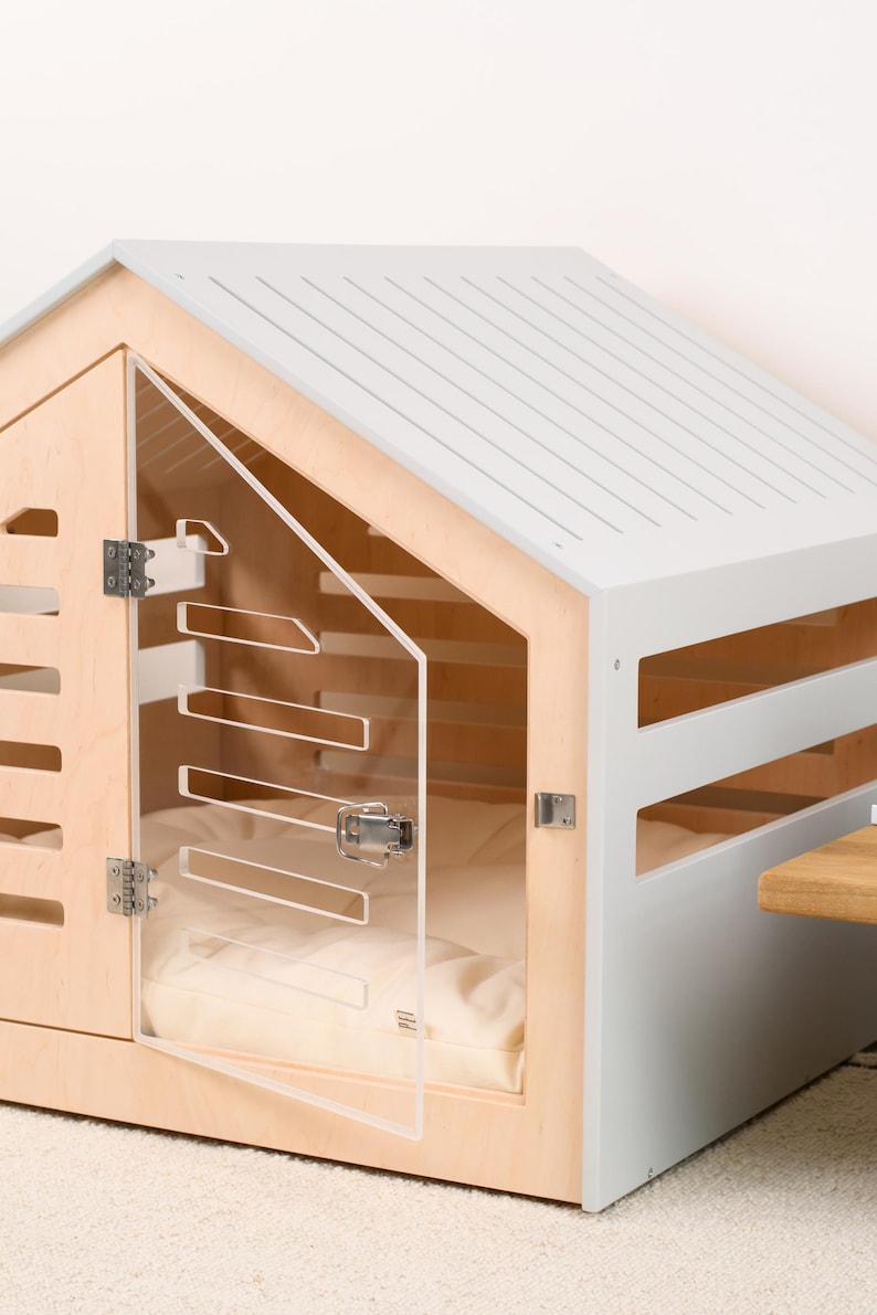 May include: A pet house featuring a light wood frame and a light gray gabled roof. The structure includes horizontal side slats and a clear acrylic door with a metal latch. Inside, a soft cream-colored cushion is visible through the transparent door.