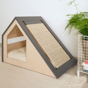 May include: A modern cat house with a built-in scratching post. The house is shaped like a small A-frame, with a light wood frame and a gray trim. The scratching post is made of woven rope. A cream-colored cushion is inside the house.