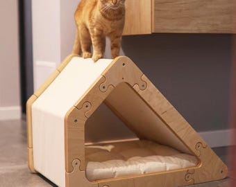 May include: A wooden cat bed shaped like a house with a white fabric roof. A ginger cat is standing on the roof of the bed.