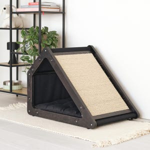 May include: A dark wooden cat house with a ramp. The house has a black interior with a black cushion. The ramp is covered in a beige woven material. The structure is placed on a white rug.
