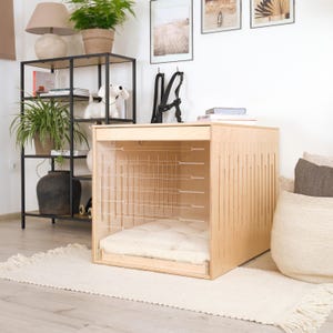 Puede incluir: Una jaula para perros de madera clara con una parte delantera de alambre y un cojín crema en el interior. La jaula tiene ranuras verticales en el lateral y está colocada sobre una alfombra blanca. Un accesorio para mascotas elegante y funcional.