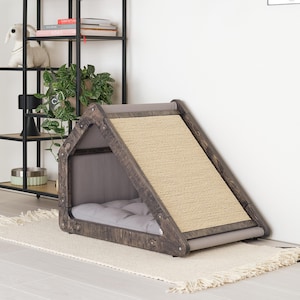 May include: A wooden pet house with a ramp and a gray cushion inside. The house is made of dark wood and has a light-colored rope scratching surface on the ramp. The house is designed for cats or small dogs.