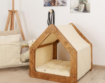 May include: A wooden dog house with a beige fabric roof and a beige cushion inside. The house is made of wood with a natural finish and has a puzzle-like design.