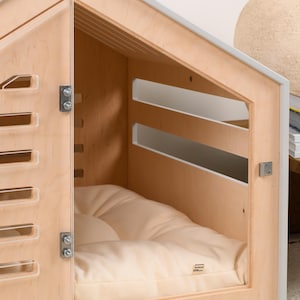 May include: A light-colored wooden pet house with a gabled roof, featuring a hinged door with horizontal slats, a matching slatted side panel, and a soft, cream-colored tufted cushion inside. The structure includes metal hinges and a small latch.