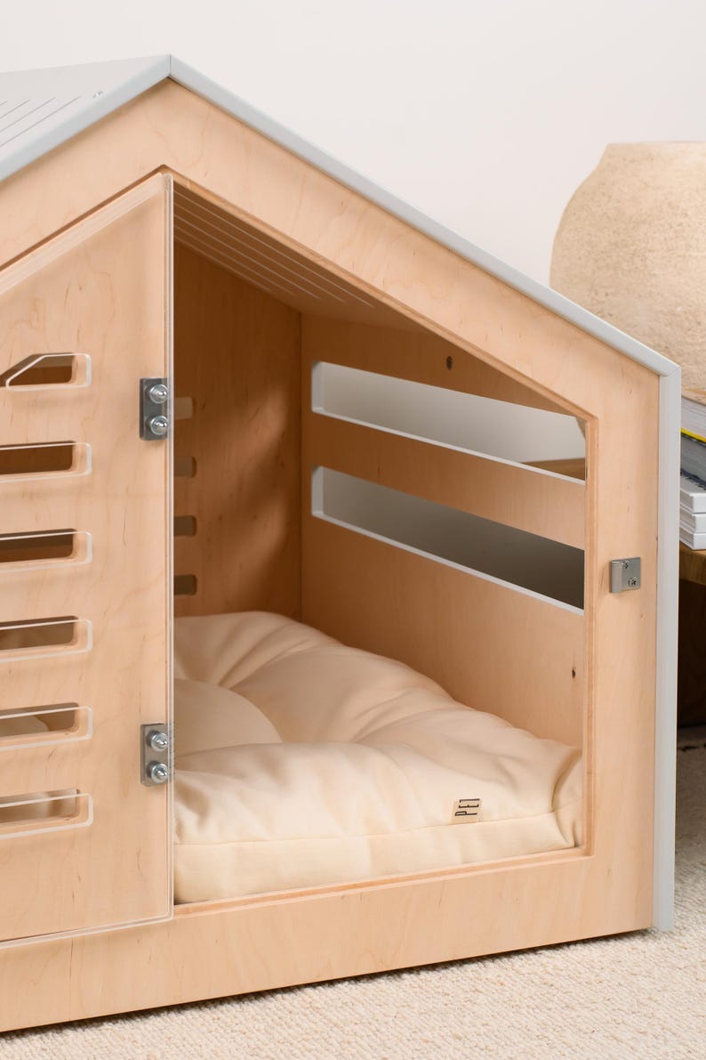May include: A light-colored wooden pet house with a gabled roof, featuring a hinged door with horizontal slats, a matching slatted side panel, and a soft, cream-colored tufted cushion inside. The structure includes metal hinges and a small latch.