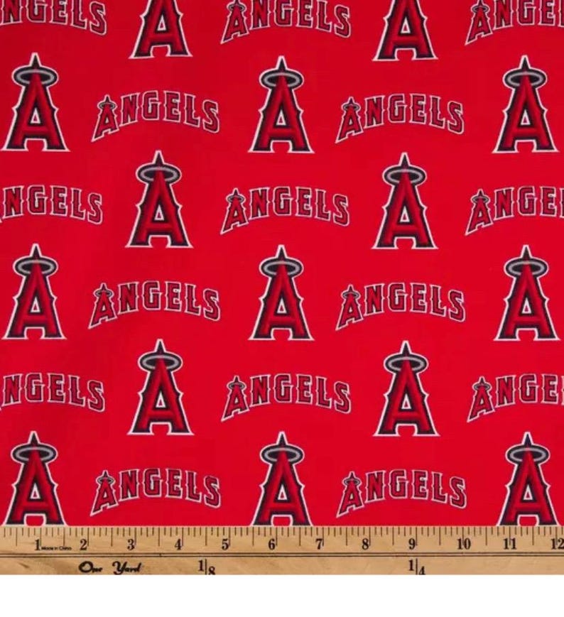 May include: Red fabric with a repeating pattern of the Los Angeles Angels baseball team logo. The logo features a stylized "A" with a halo and the word "ANGELS" in white. A ruler is at the bottom, measuring up to 12 inches.