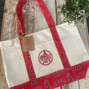 May include: Cream-colored tote bag with red accents featuring the text "ANGELS" and the team logo. The bag has a red trim and handles with the team's logo. A tag reads "TRADER JOE'S".