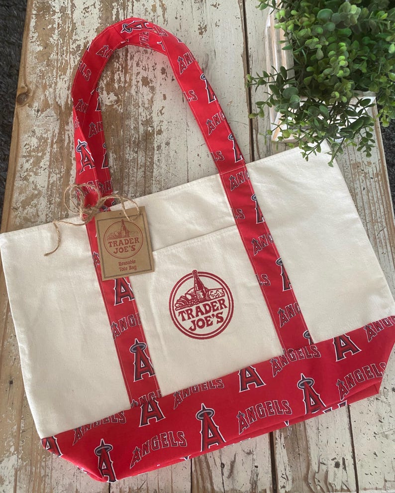 May include: Cream-colored tote bag with red accents featuring the text "ANGELS" and the team logo. The bag has a red trim and handles with the team's logo. A tag reads "TRADER JOE'S".