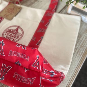 May include: Cream-colored canvas tote bag with red accents. The bag features red straps and a red fabric base printed with the word "ANGELS" and the letter "A". The Trader Joe's logo is visible on the front of the bag.