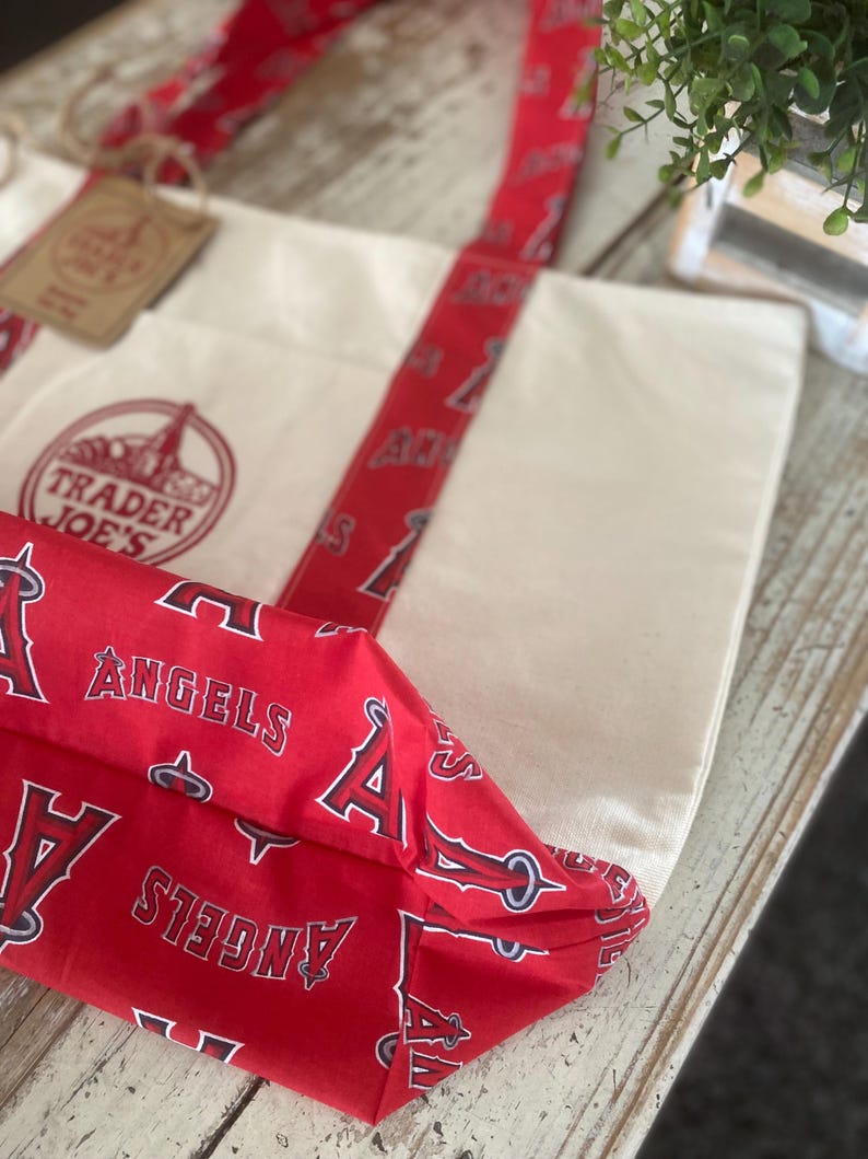 May include: Cream-colored canvas tote bag with red accents. The bag features red straps and a red fabric base printed with the word "ANGELS" and the letter "A". The Trader Joe's logo is visible on the front of the bag.