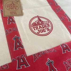 May include: Cream-colored reusable tote bag with red accents. The bag features a red and white design with the text "ANGELS" and the letter "A". The Trader Joe's logo is printed on the front.