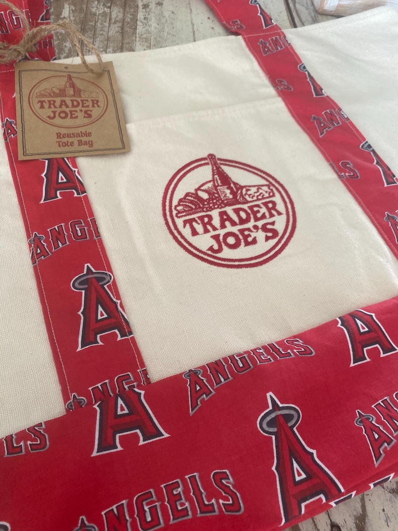 May include: Cream-colored reusable tote bag with red accents. The bag features a red and white design with the text "ANGELS" and the letter "A". The Trader Joe's logo is printed on the front.