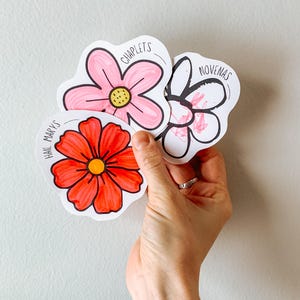 May include: Four flower-shaped paper cutouts with handwritten text and floral illustrations. The flowers are in shades of pink and red, with yellow centres. The text includes "CHAPLETS", "NOVENAS", and "HAIL MARYS".