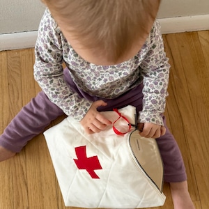 Op de afbeelding: Een witte, gewatteerde tas met een rode kruisapplicatie. De tas heeft een zwarte rits en een rood lint. De tas ligt op een houten vloer.