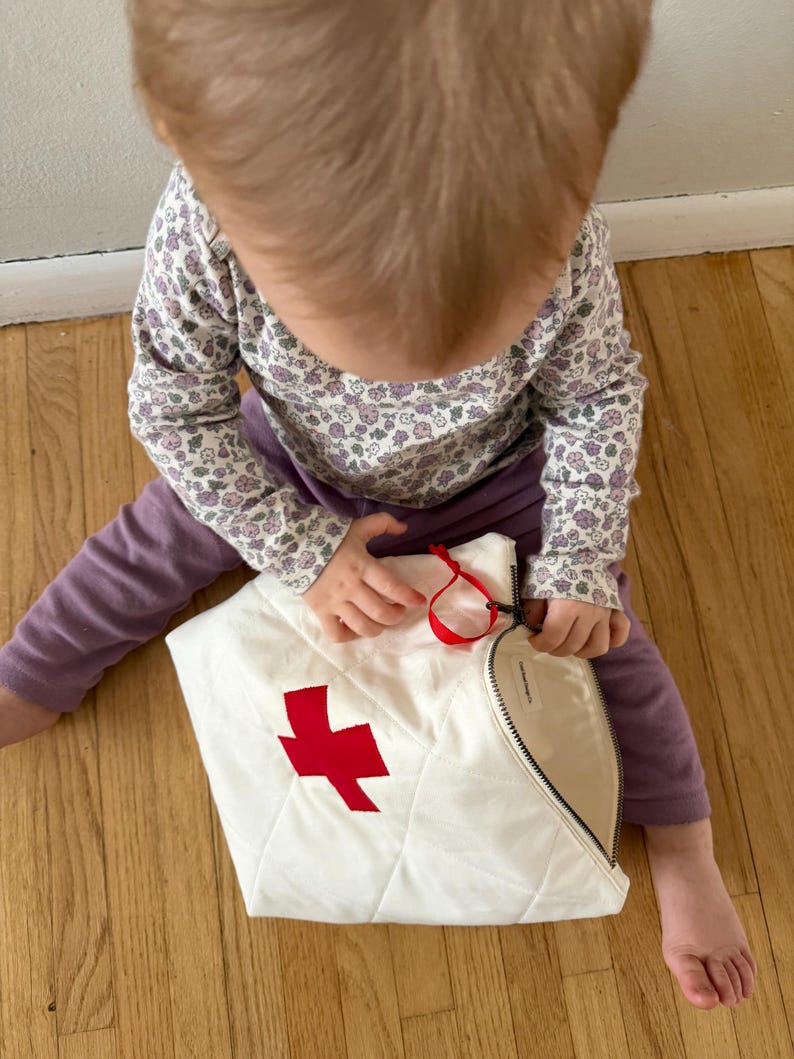 Op de afbeelding: Een witte, gewatteerde tas met een rode kruisapplicatie. De tas heeft een zwarte rits en een rood lint. De tas ligt op een houten vloer.