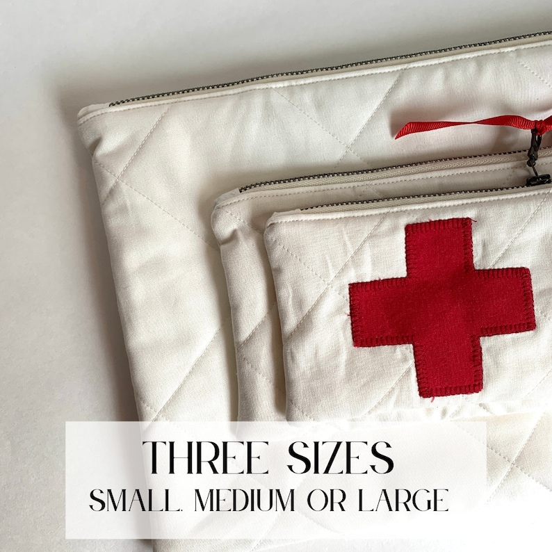 Op de afbeelding: Drie witte gewatteerde ritszakjes in verschillende maten. Het kleinste zakje heeft een grote rode kruisapplicatie. De tekst "THREE SIZES SMALL MEDIUM OR LARGE" wordt weergegeven in een witte rechthoek.