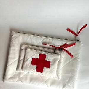 Op de afbeelding: Drie witte gewatteerde etuis van verschillende formaten, genest in elkaar. Elke etui heeft een zilveren ritssluiting en een rood lint. De kleinste etui heeft een rode kruisapplicatie, wat wijst op een medisch thema.