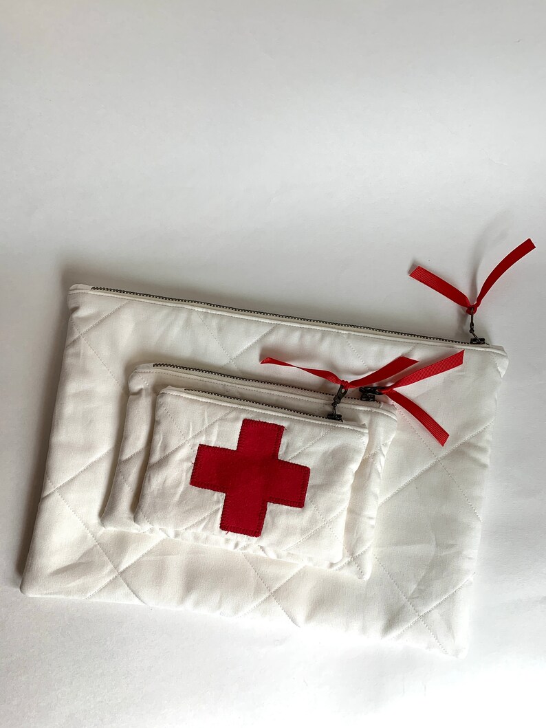 Op de afbeelding: Drie witte gewatteerde etuis van verschillende formaten, genest in elkaar. Elke etui heeft een zilveren ritssluiting en een rood lint. De kleinste etui heeft een rode kruisapplicatie, wat wijst op een medisch thema.
