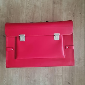 Puede incluir: Un maletín de cuero rojo brillante con forma rectangular. Tiene un cierre de solapa con dos cierres metálicos plateados y un bolsillo delantero más pequeño. El maletín está sobre una superficie de madera.